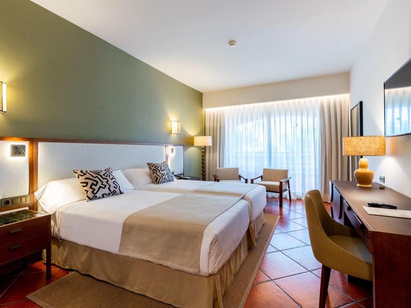 Gemütliches Hotelzimmer mit Doppelbett, Schreibtisch, Stühlen und großem Fenster mit Vorhängen.