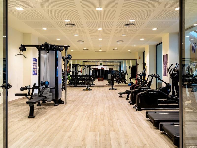 Heller Fitnessraum mit Holzboden, Kraft- und Cardiogeräten sowie großem Spiegel an der Rückwand.