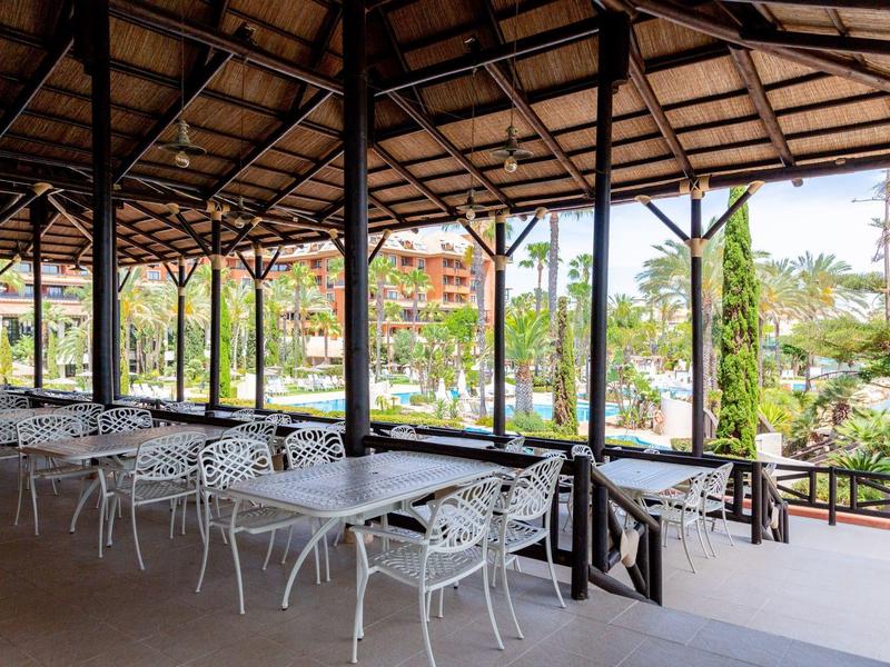 Überdachte Terrasse mit weißen Metallstühlen und Tischen, Blick auf Grünflächen und Gebäude.