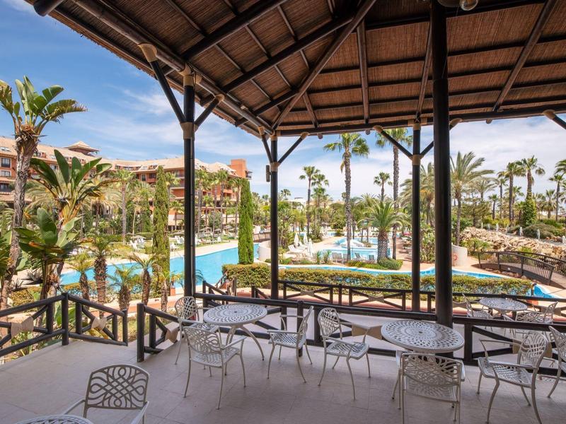 Überdachte Terrasse mit weißen Tischen und Stühlen, Blick auf Pool, Palmen und Hotelgebäude.