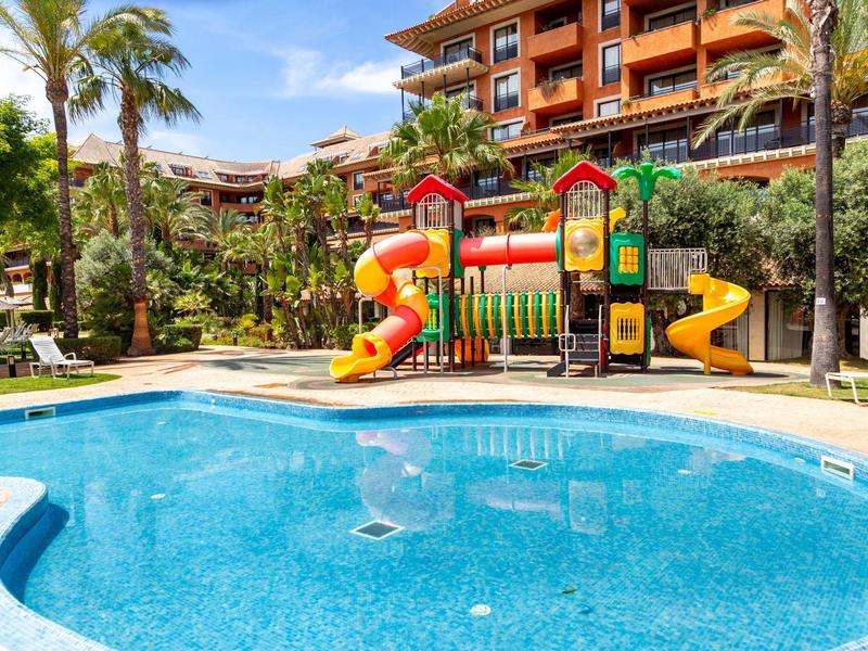 Kinderspielplatz mit bunten Rutschen neben klarem, rundem Pool und Palmen unter blauem Himmel.