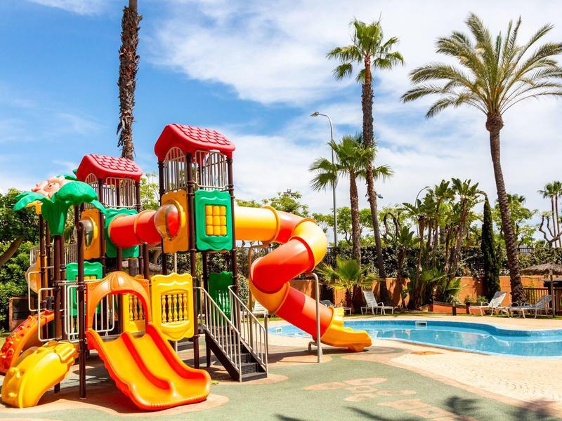 Bunter Kinderspielplatz mit Rutschen neben einem Pool, umgeben von Palmen und blauen Himmel.