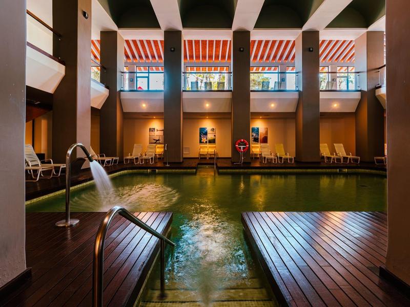 Innenraum mit beheiztem Schwimmbecken, Holzboden, großen Fenstern und gestreiften Markisen.