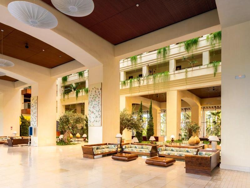 Großes offenes Hotel-Lobby mit beigen Wänden, Holzdecke, Pflanzen und braunen Sofas.