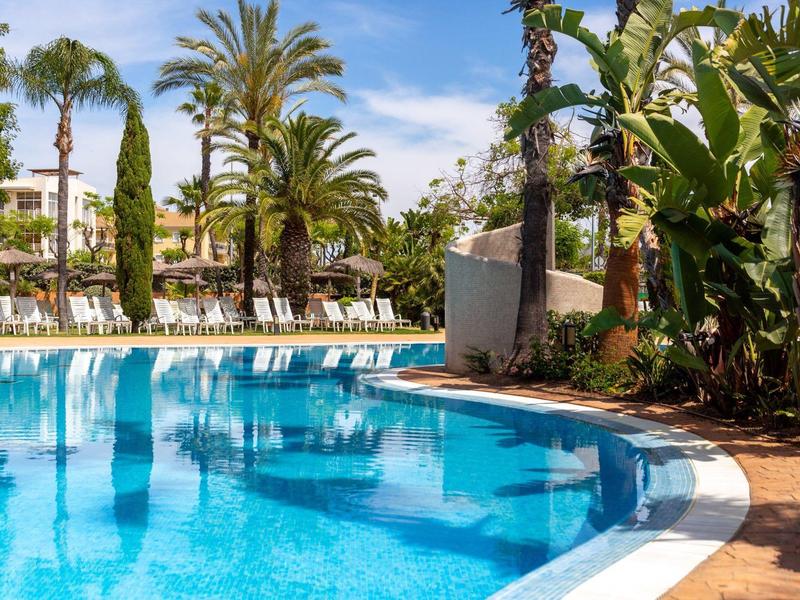 Großer Pool mit blauem Wasser, umgeben von Palmen und Liegestühlen unter blauem Himmel.