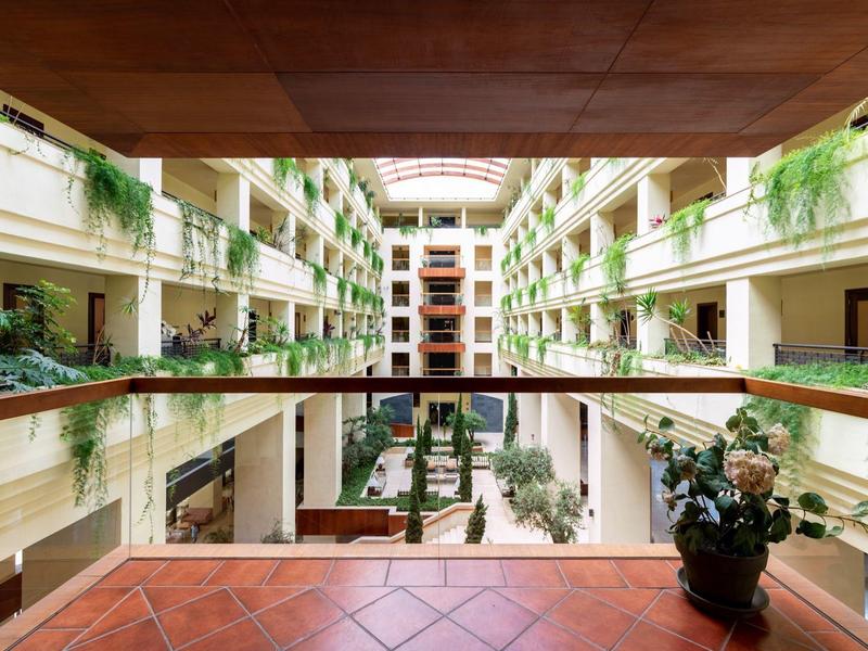 Großes Atrium mit Glasdach, begrünte Balkone und Fliesenboden, Pflanze im Vordergrund.