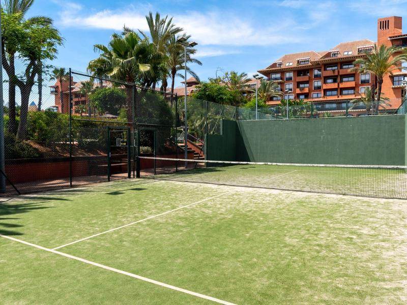 Padel-Tennisplatz mit grünem Kunstrasen, umgeben von Palmen und Gebäuden im Hintergrund.