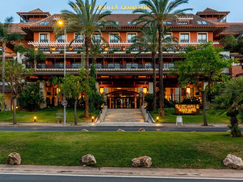 Großes Hotel mit roten Dächern, Palmen und beleuchtetem Eingangsbereich bei Dämmerung.