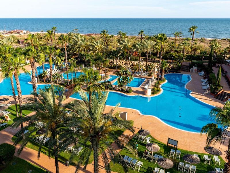 Großer blauer Pool mit Palmen und Liegestühlen vor dem Meer unter klarem Himmel.