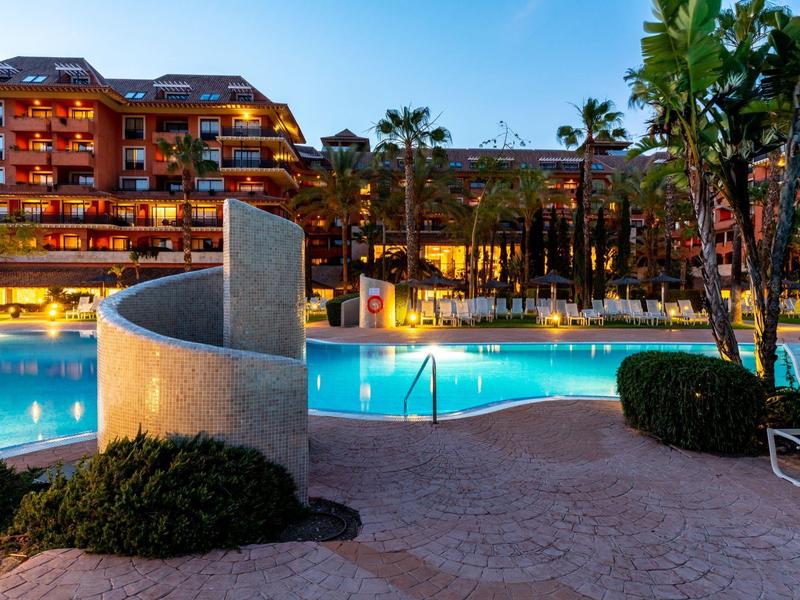 Ein beleuchteter Hotelpool bei Dämmerung, umgeben von Palmen und roten Gebäuden.