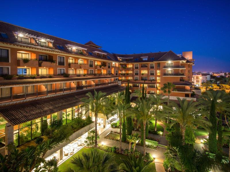 Großes modernes Hotel in warmem Licht mit Balkonen, umgeben von grünen Palmen und blauem Abendhimmel.