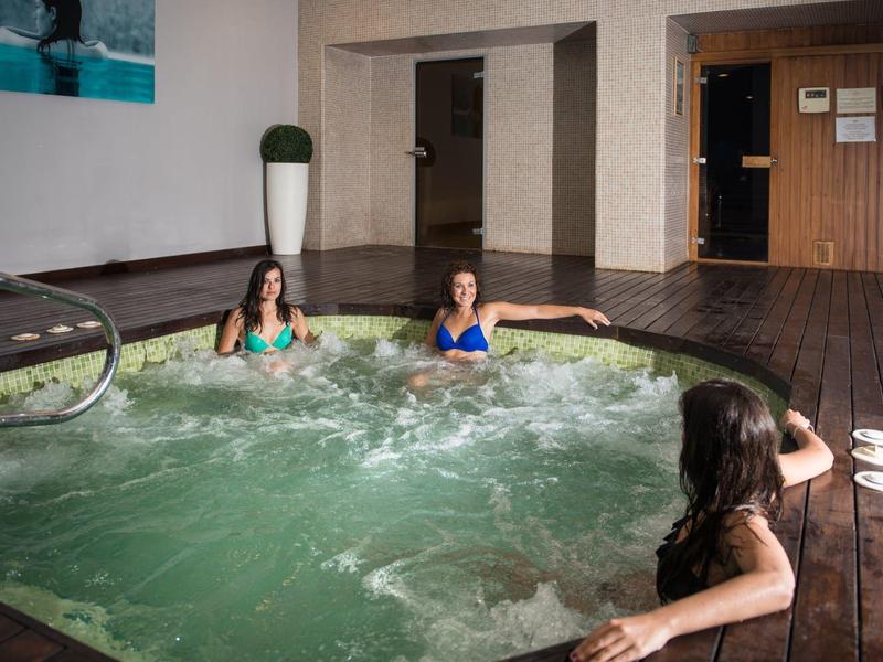 Drei Frauen entspannen im sprudelnden Hallenbad mit Holzdeck und heller Wandgestaltung.