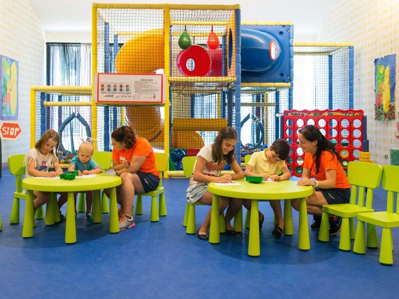 Kinder spielen an grünen Tischen mit buntem Klettergerüst und blauem Bodenbelag in hellem Raum.