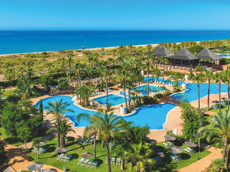 Große Resortanlage mit mehreren blauen Pools, Palmen und Blick auf das blaue Meer am Horizont.