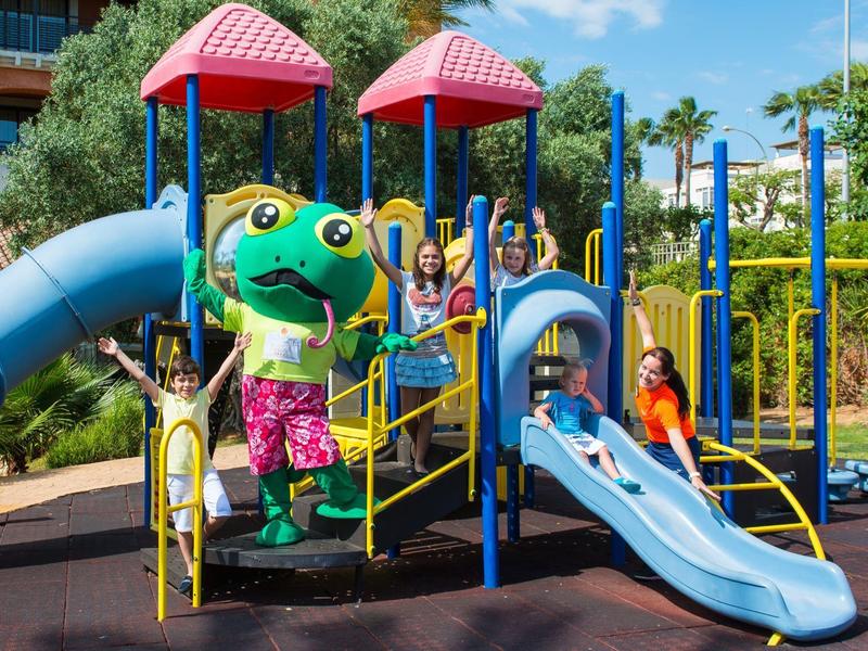 Bunter Spielplatz mit Rutsche, Kindern und großem grünen Frosch-Maskottchen bei Sonnenschein.