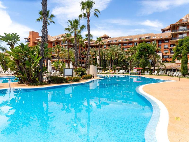 Großer, blauer Pool mit Palmen und rotem Hotelgebäude unter blauem Himmel.