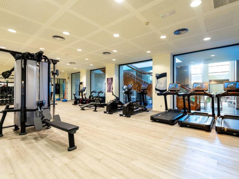 Moderner Fitnessraum mit Laufbändern, Kraftgeräten und großen Fenstern, heller Holzboden.