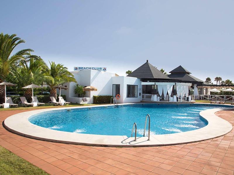 Runder blauer Pool mit weißem Rand und Palmen vor weißem Gebäude unter blauem Himmel.