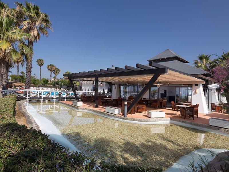Modernes Gebäude mit großer Terrasse, umgeben von Palmen und klarem Wasserbecken bei blauem Himmel.