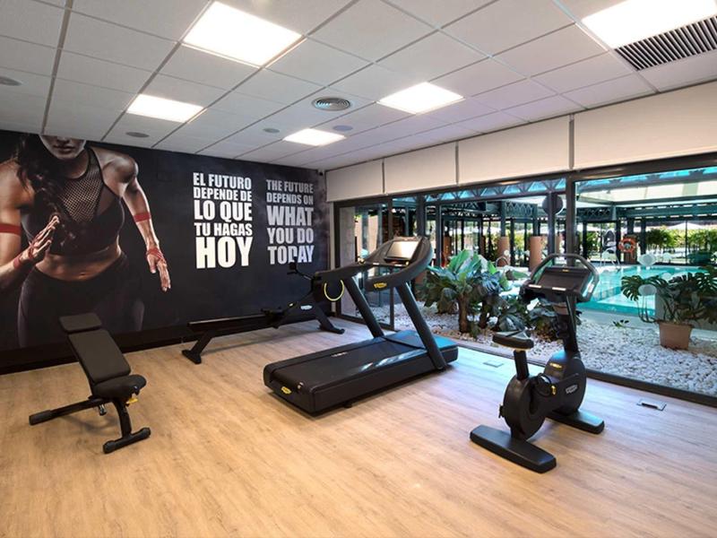 Moderner Fitnessraum mit Laufband, Trainingsrad, Hantelbank und großem Fenster zum Außenbereich.