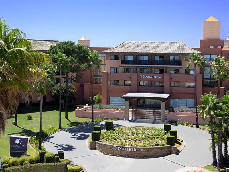 Mehrstöckiges Gebäude mit roter Fassade, umgeben von Palmen und gepflegtem Grün.