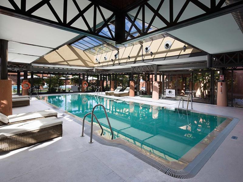 Innenraum mit großem Schwimmbecken, Glasdach, weißen Liegen und moderner Architektur.