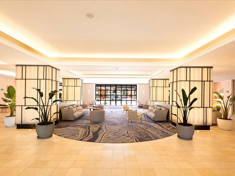 Heller, großzügiger Raum mit runden Teppichen, vielen Pflanzen und warmem Deckenlicht.