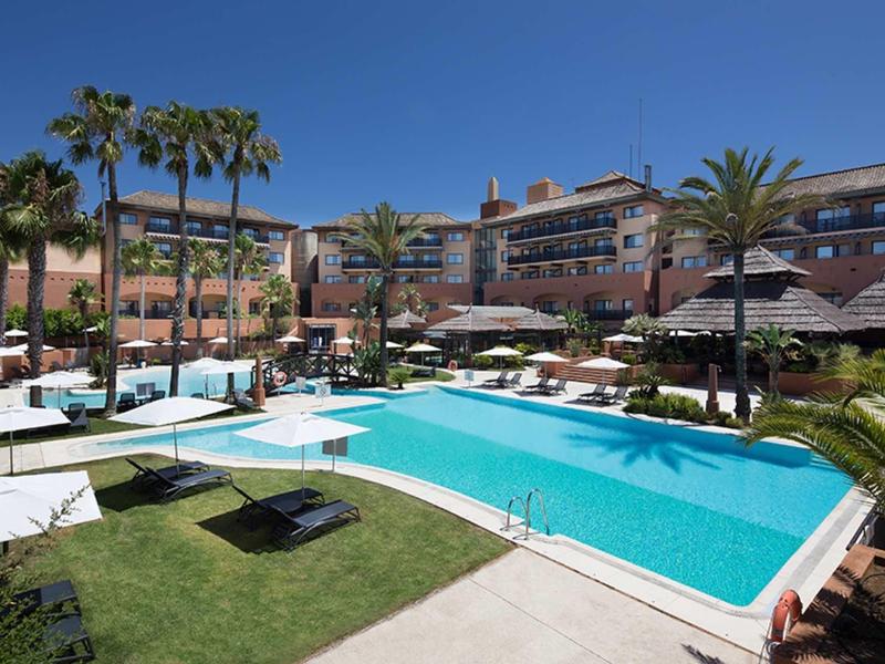 Großer rechteckiger Pool mit Liegen, Palmen und Gebäuden im Hintergrund bei sonnigem Wetter.