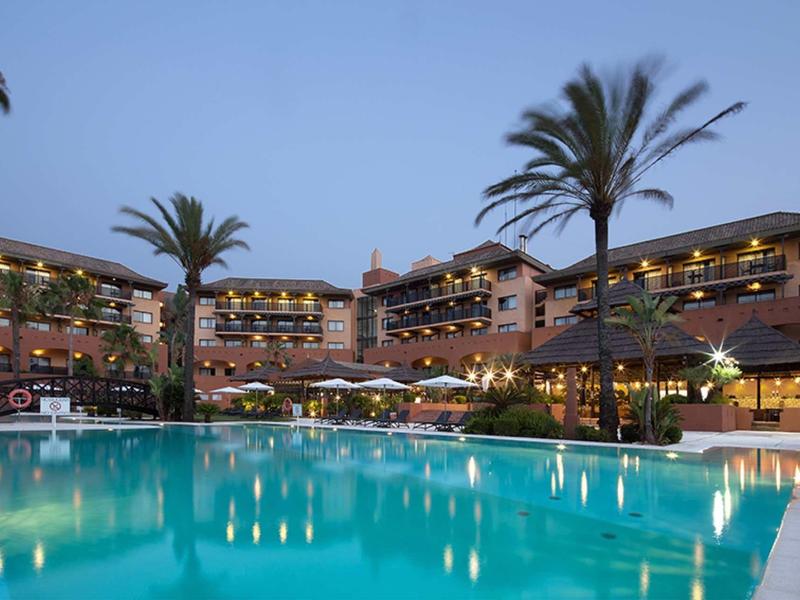 Großes Hotel mit beleuchtetem Pool und hohen Palmen bei klarem Abendhimmel.