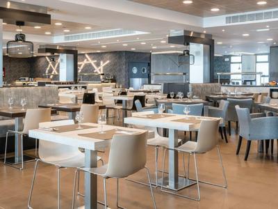 Moderner, heller Speiseraum mit weißen und grauen Stühlen, quadratischen Tischen und großen Fenstern.