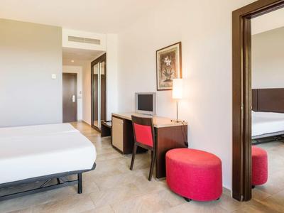 Camera d'hotel moderna con due letti, grande specchio, scrivania con sedia e sgabello rosso.