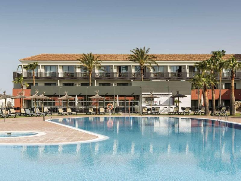 Großer Außenpool mit klar blauem Wasser vor einem mehrstöckigen Hotelgebäude und Palmen.