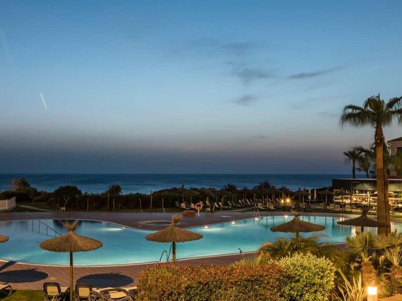 Moderner Hotelpool bei Dämmerung mit Palmen und Blick auf ruhiges Meer unter blauem Himmel.
