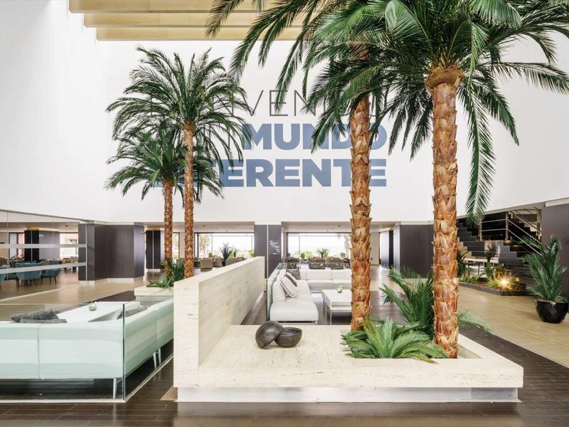 Moderner Innenraum mit Palmen, Sitzbereichen und hellem, luftigen Design mit viel Tageslicht.