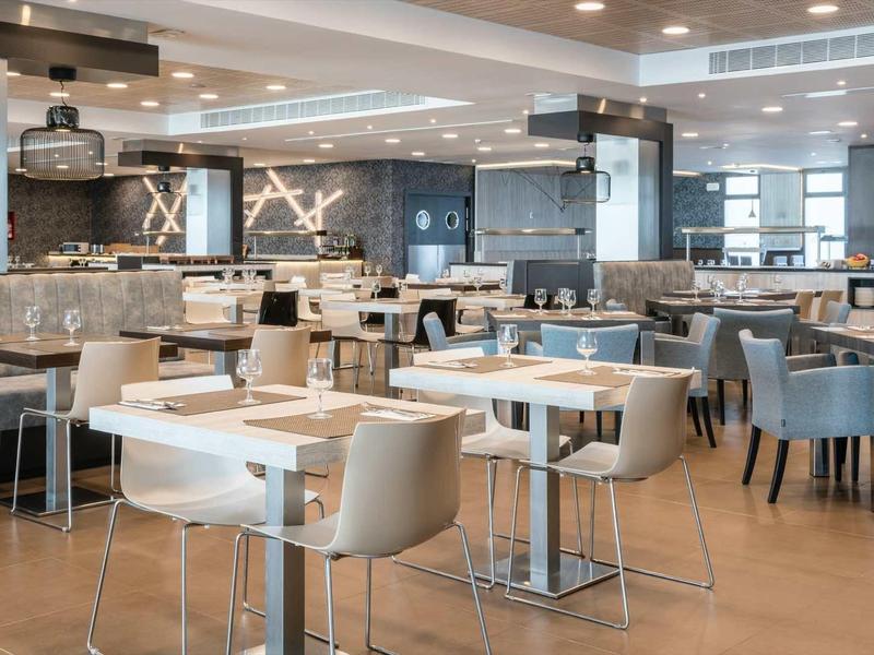 Moderner, heller Speiseraum mit weißen und grauen Stühlen, quadratischen Tischen und großen Fenstern.