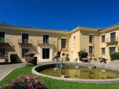 Beiger Gebäudekomplex mit zwei Etagen um kreisrunden Springbrunnen und grüner Wiese.