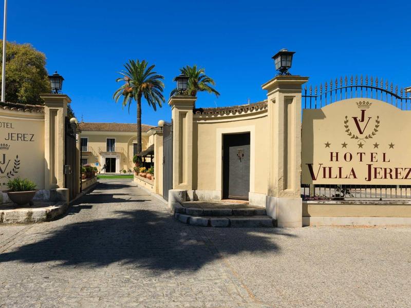 Eingang zum Hotel Villa Jerez mit hohen Säulen, Palmen und sonnigem, blauem Himmel.
