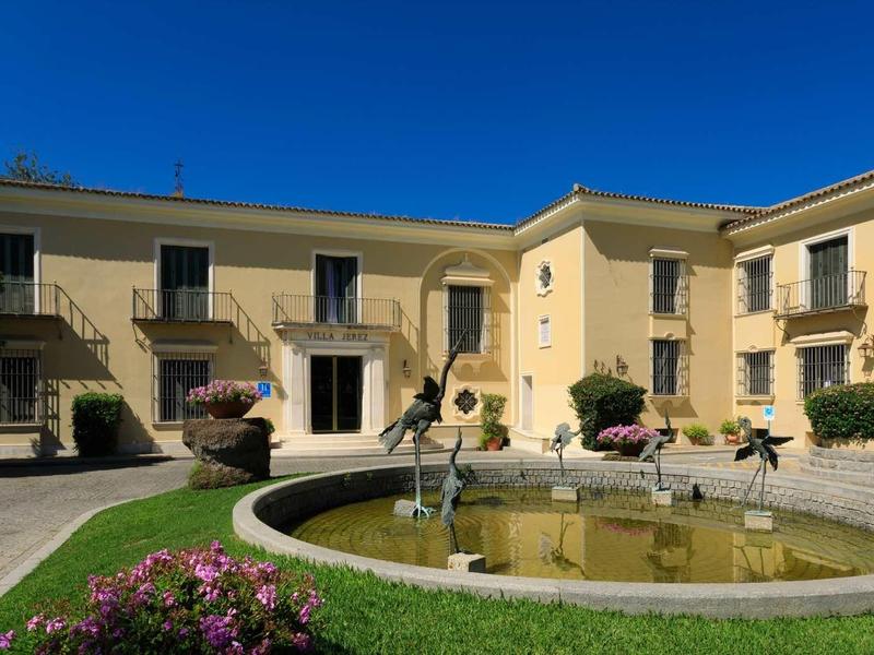 Beiger Gebäudekomplex mit zwei Etagen um kreisrunden Springbrunnen und grüner Wiese.