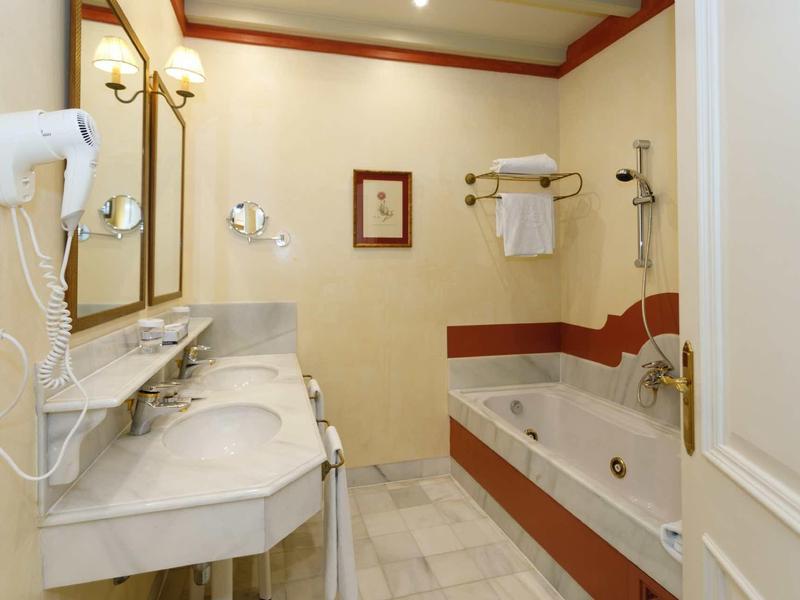 Helles Badezimmer mit Doppelwaschbecken, Badewanne, Handtüchern und Haartrockner an der Wand.