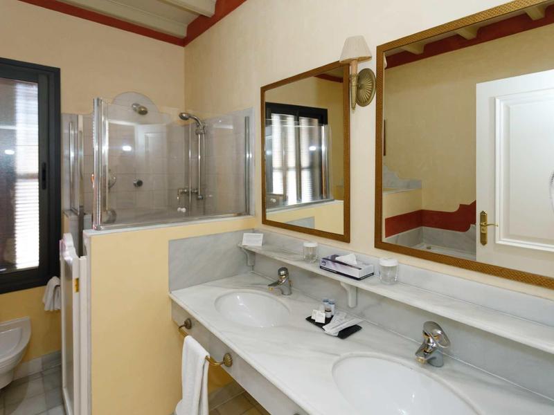 Helles Badezimmer mit Doppelwaschbecken, großer Spiegel, Glasdusche und Fenster mit Holzrahmen.