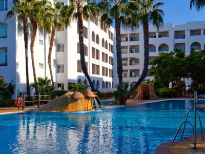 Edificio alberghiero a più piani circondato da palme e grande piscina sotto un cielo azzurro.
