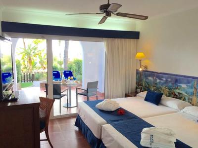 Camera d'hotel luminosa con letto matrimoniale, ventilatore a soffitto e terrazza con vista su giardino tropicale.