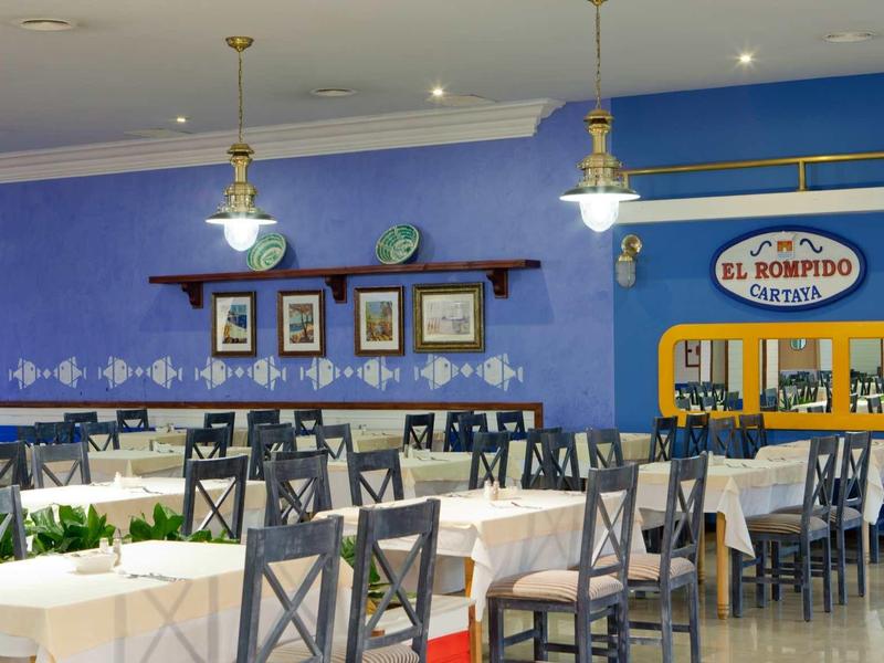 Restaurant mit weißen Tischdecken, blauer Wand und zahlreichen Stühlen und Tischen.