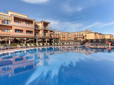 Großes Hotel mit mehreren Stockwerken, umgeben von Sonnenschirmen, Pools und blauem Himmel.