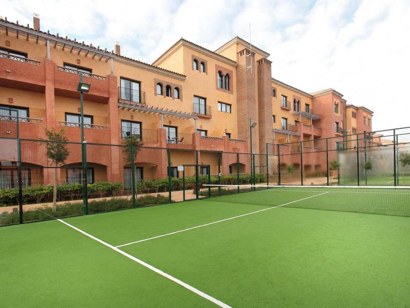 Grüner Tennisplatz mit klaren Linien vor orangefarbenen Wohngebäuden unter blauem Himmel.