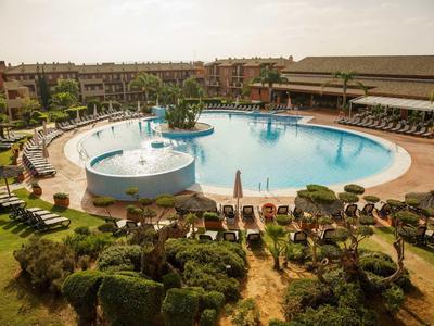 Großer Rundpool mit Sonnenliegen, Sonnenschirmen und grünen Büschen rundum bei sonnigem Wetter.