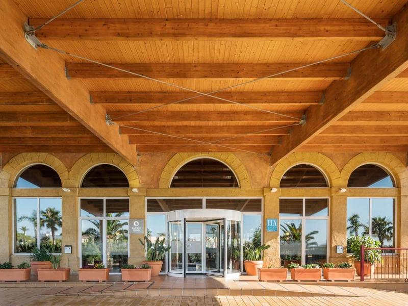 Eingang mit großen Glasfenstern und -türen, darüber Holzdecke mit Balken und Pflanzen vor den Fenstern.