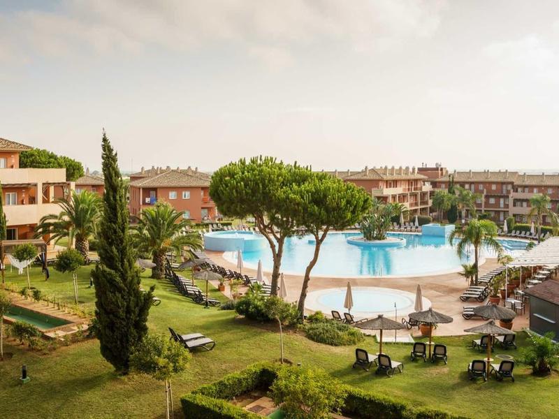 Große Hotelanlage mit Pool, Liegestühlen, Palmen und gepflegtem Grün unter bewölktem Himmel.