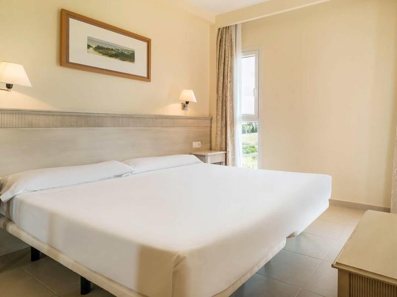 Helles Schlafzimmer mit Doppelbett, weißen Wänden, Holzmöbeln und Fenster mit Vorhängen.