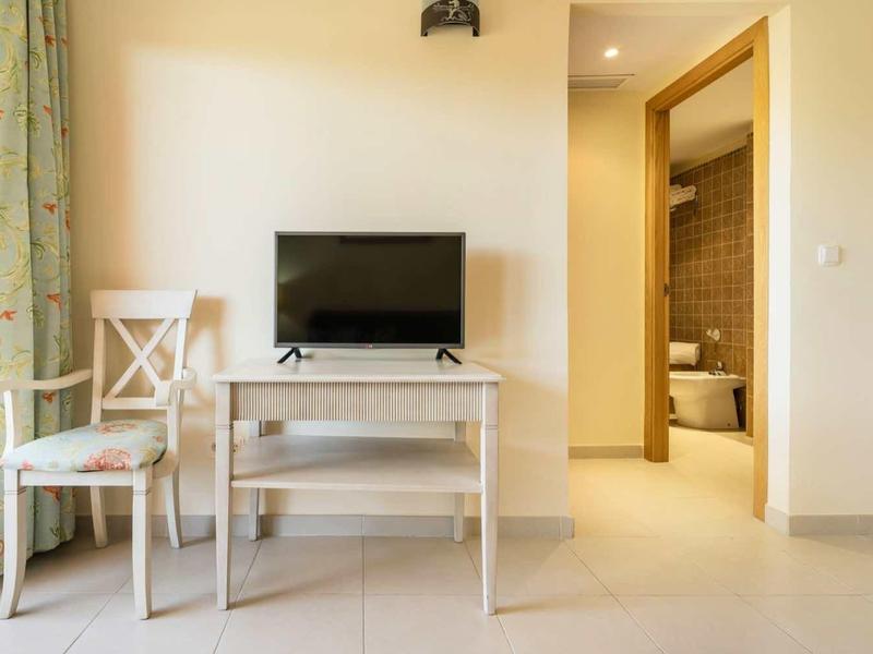 Helles Wohnzimmer mit Flachbildfernseher, Glastisch, Holzstühlen und Zugang zu Badezimmer und Balkon.